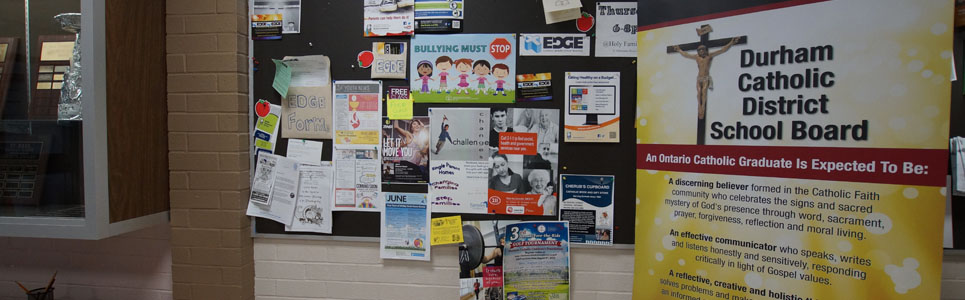Stand-up sign stating the Catholic Graduate Expectations beside a bulletin board with various school-related flyers.